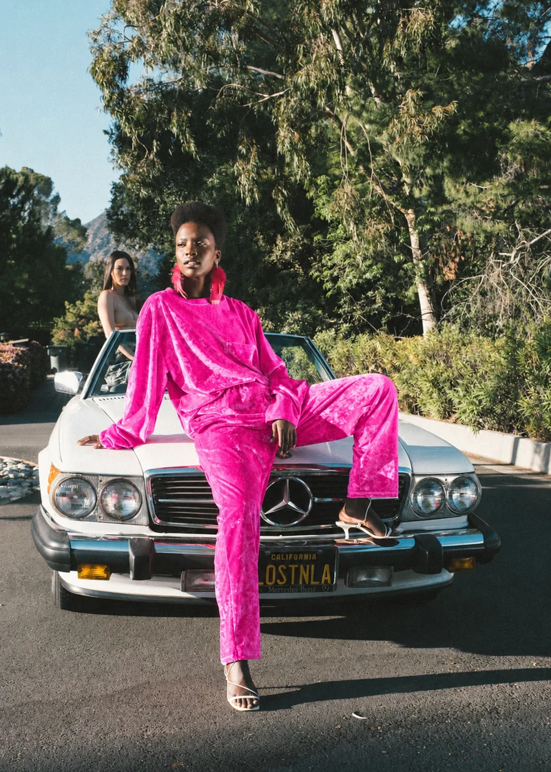 Model in hot-pink crushed velvet seated on a white Mercedes, with full outfit and shoes visible