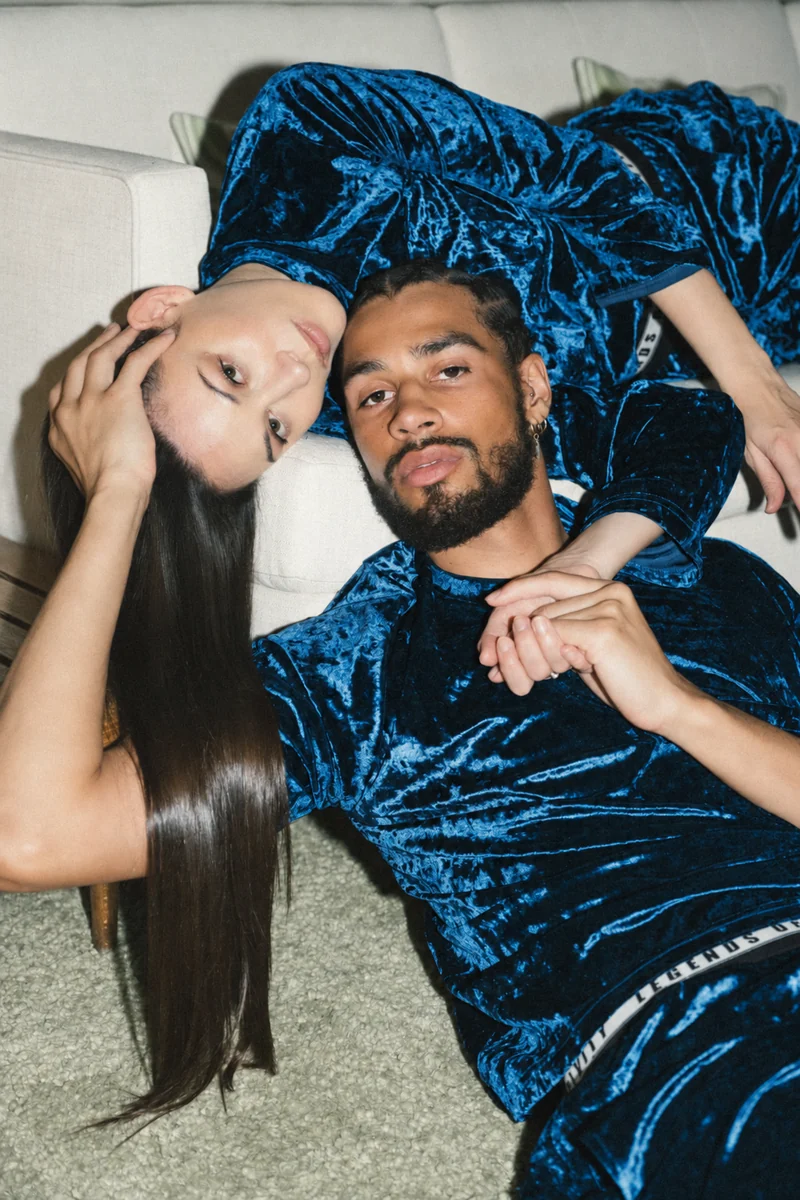 Two models reclining together beside a cream sofa in matching blue crushed-velvet sets
